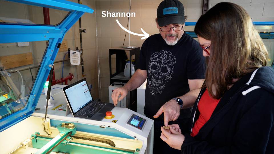 a light skinned man with a grey beard and a black shirt and baseball hat shoes a younger woman how to use the laser machine.