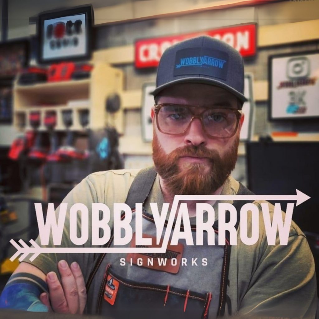 a man with a baseball hat and a red ginger beard and sunglasses in in a garage. the words wobbly arrow signworks are overliad