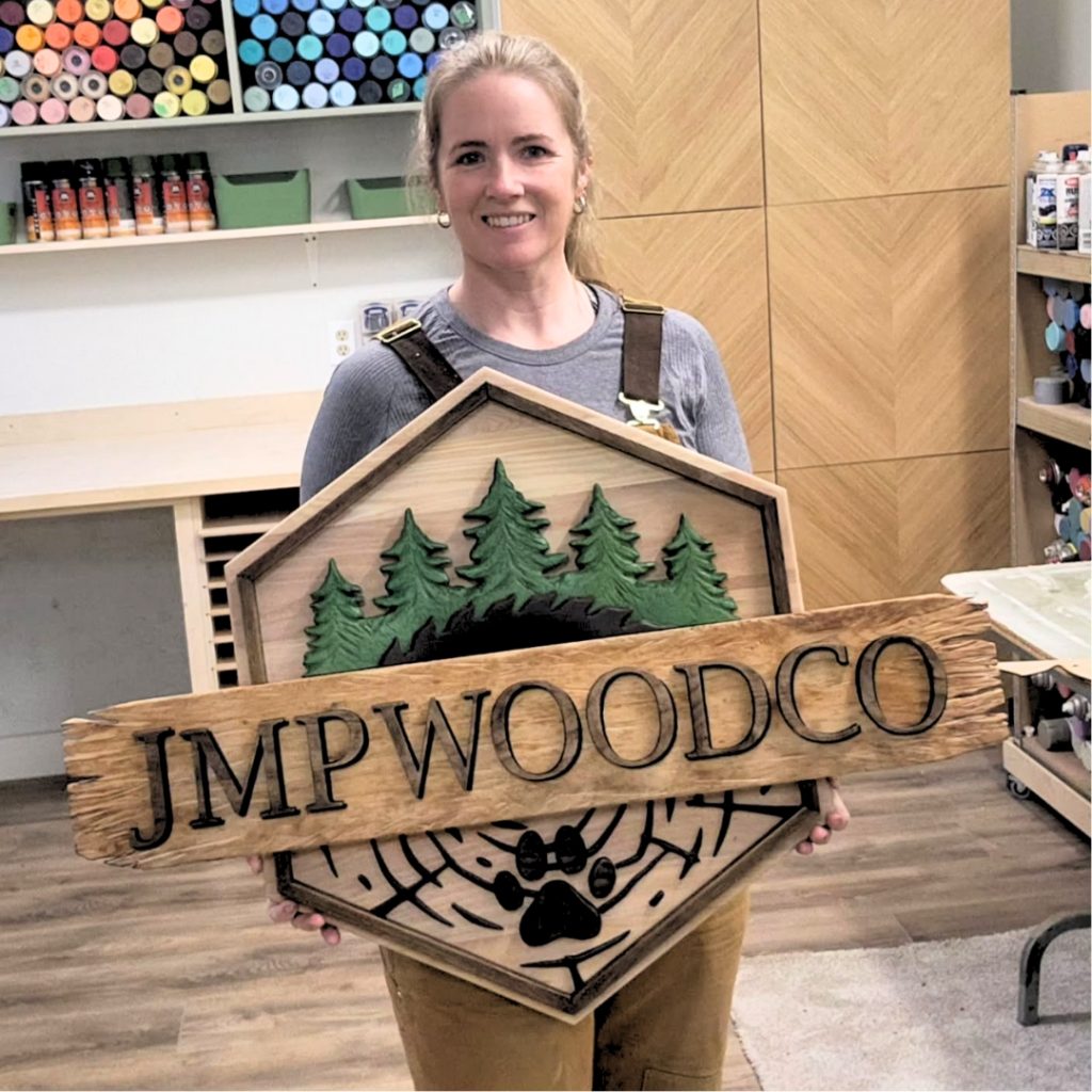 a light skinned woman (Jenny)with her hair in a pontytail is wearing coveralls holding a wood sign that says JMP wood co