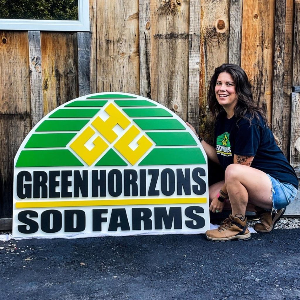 a woman Jess Seaton, kneels next to a laser cut sign that says Green horizon Sod farms