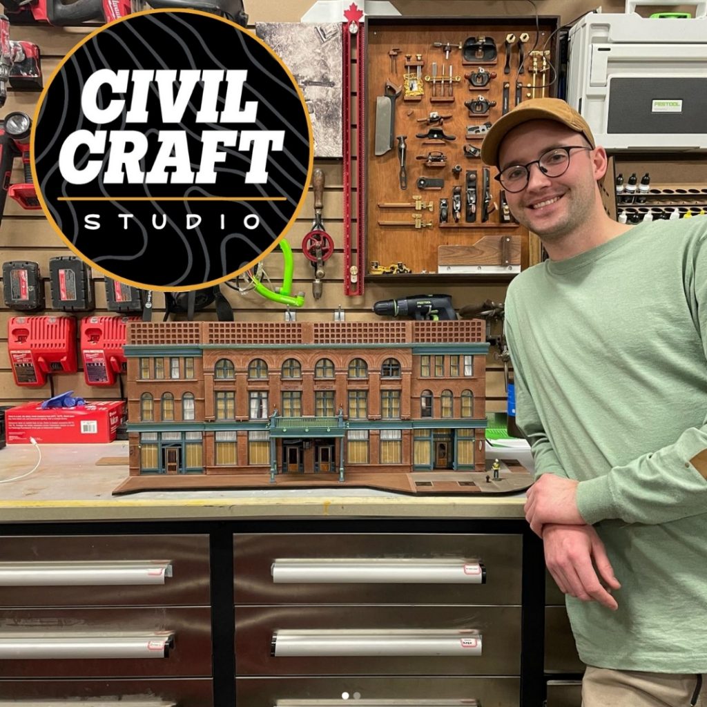 Ryan a young man with glasses sits leaning on a desk with a miniature model of a hotel.