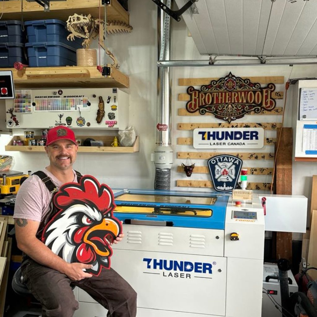 A man with a moustache holds a sign of a cartoon angry chicken head sitting in his garage infront of his blue and white metal thunder laser cutter machine