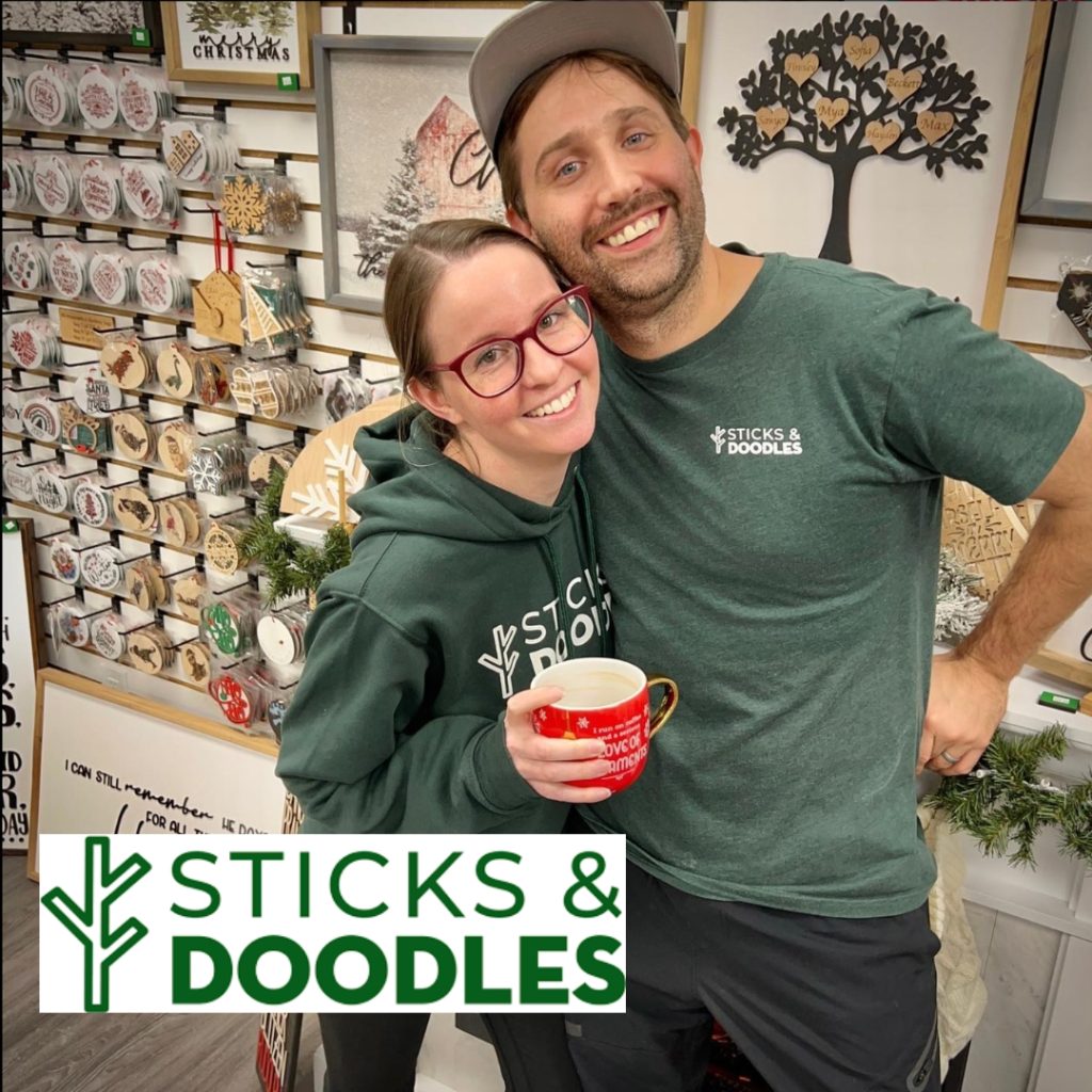 a young woman and man hug infront of a display of oranments made on the laser cutter. they are wearing dark green shirts with their company logo